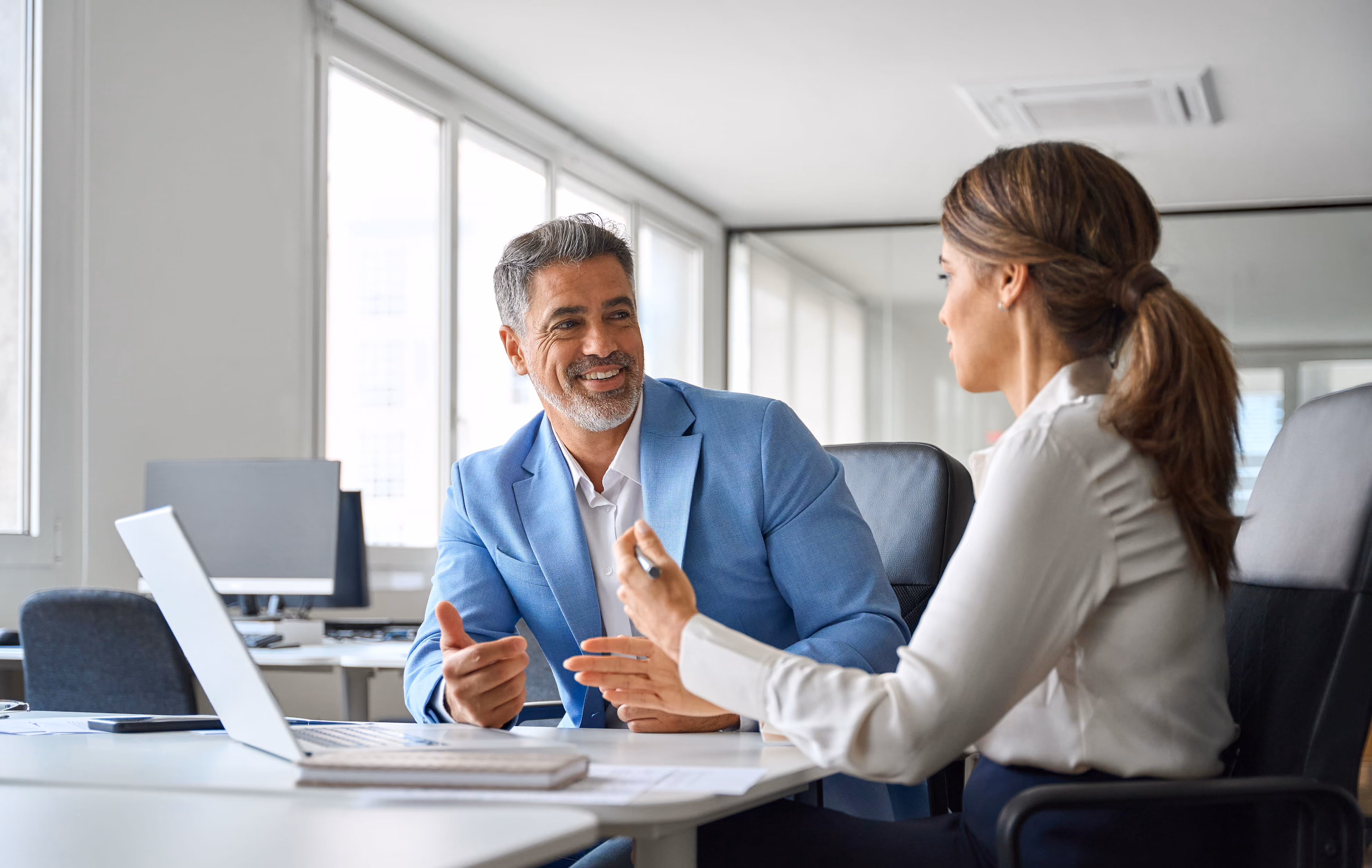 Smiling executive in a light blue blazer meeting with a colleague at an office desk