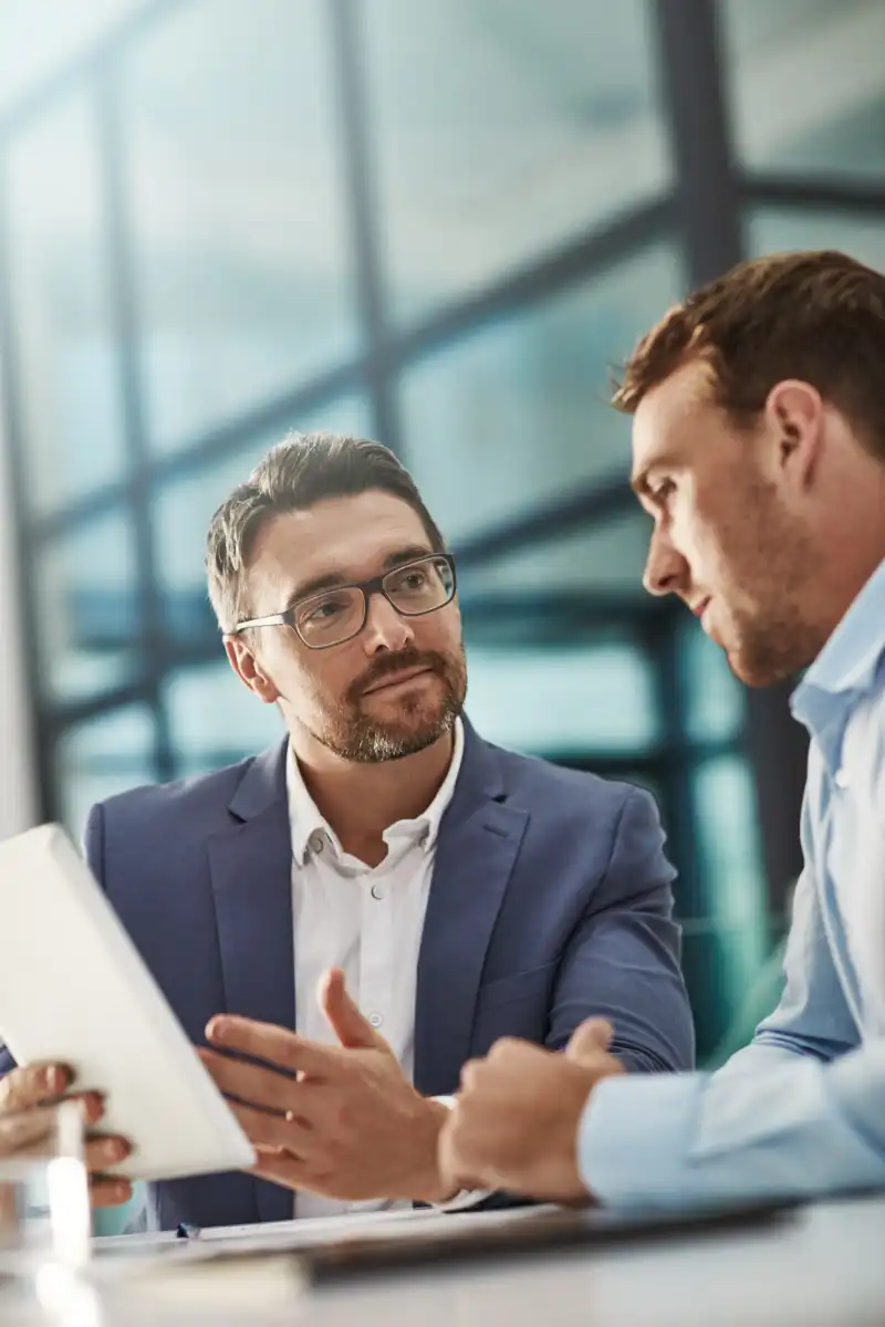 Two businessmen discussing plans on a tablet in a modern glass-walled office