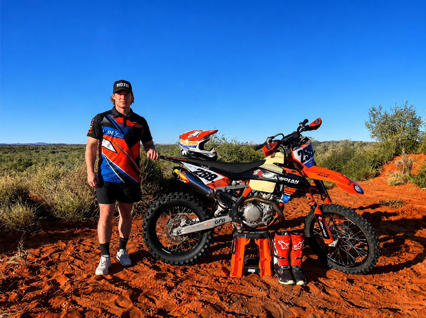 Man in motocross gear standing beside orange dirt bike with helmet and boots on red dirt ground.
