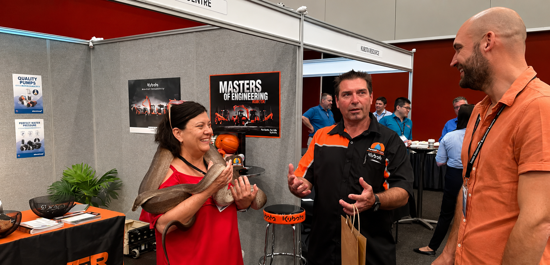 A woman holds a large snake around her shoulders while two men talk and smile at her at a booth.
