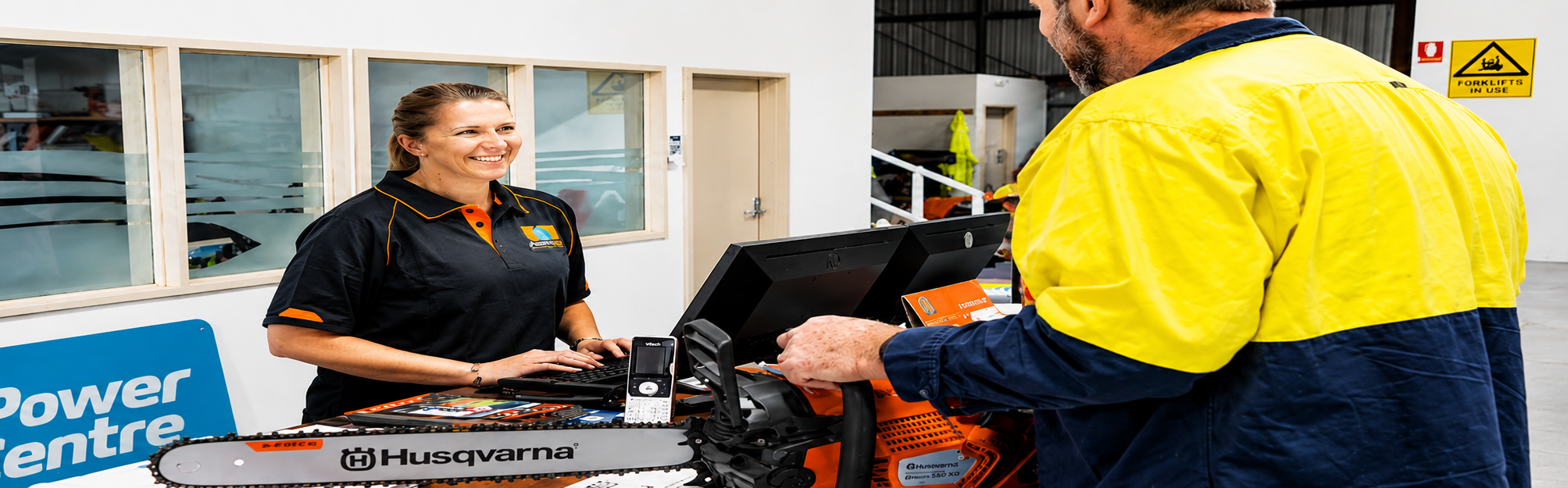 Smiling store employee helps customer holding a Husqvarna chainsaw at service counter.