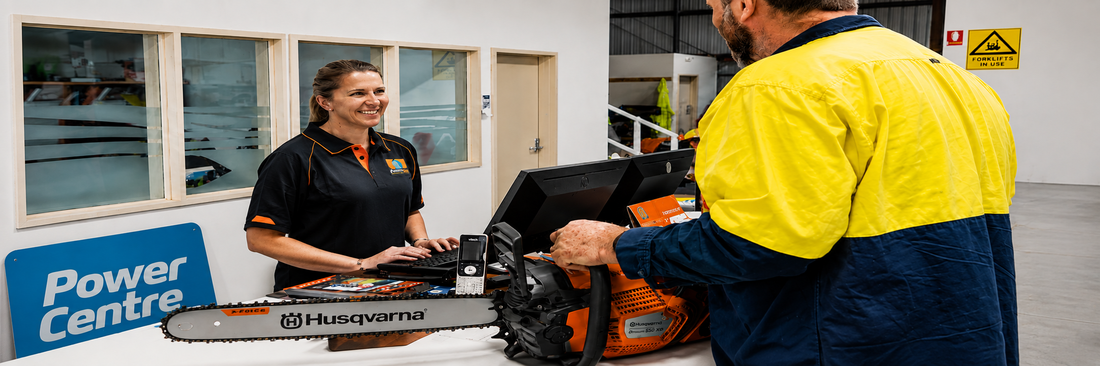 Smiling store employee helps customer holding a Husqvarna chainsaw at service counter.