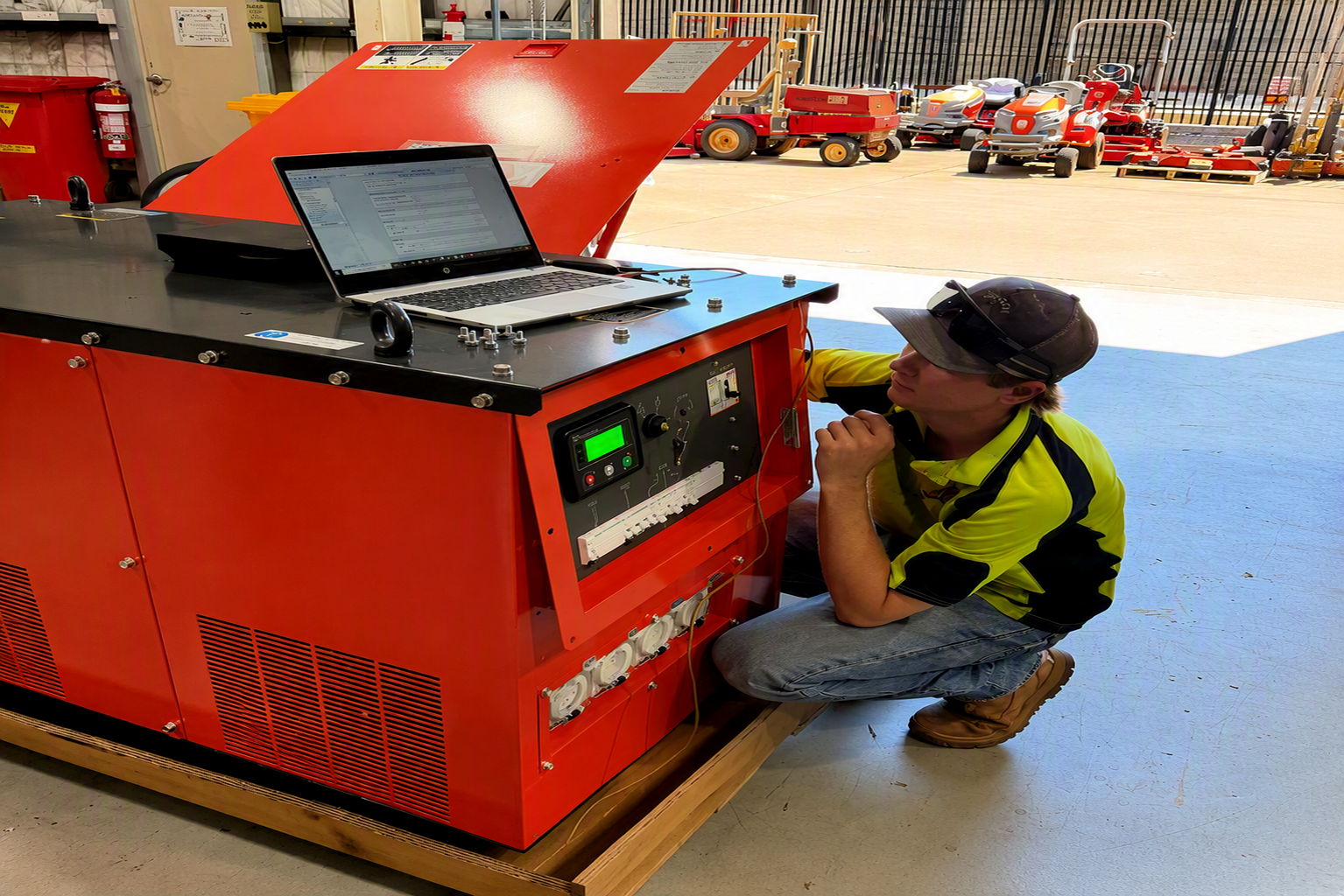 Technician in yellow shirt and cap inspecting red industrial generator with laptop on top in workshop.
