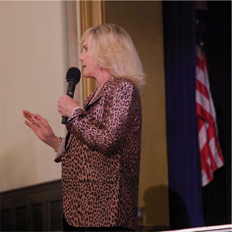 Erin Brockovich speaks into a microphone on stage, with a blurred American flag in the background.