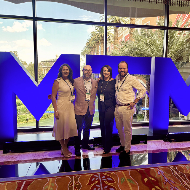 Four people smiling in front of large blue letters "MTM" with a window view of palm trees and a sunny sky in the background.