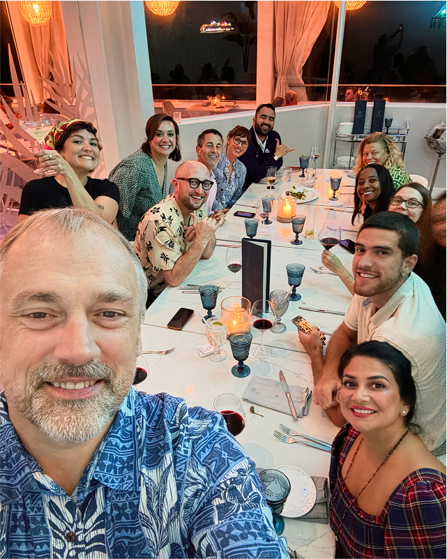 A group of people smiling around a dinner table, with plates, glasses, and candles. A man in the foreground takes a selfie.