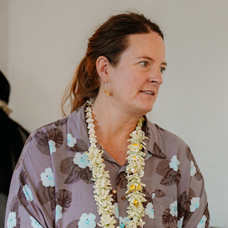 Woman wearing a floral shirt and a lei, looking to the side, standing indoors against a plain background.
