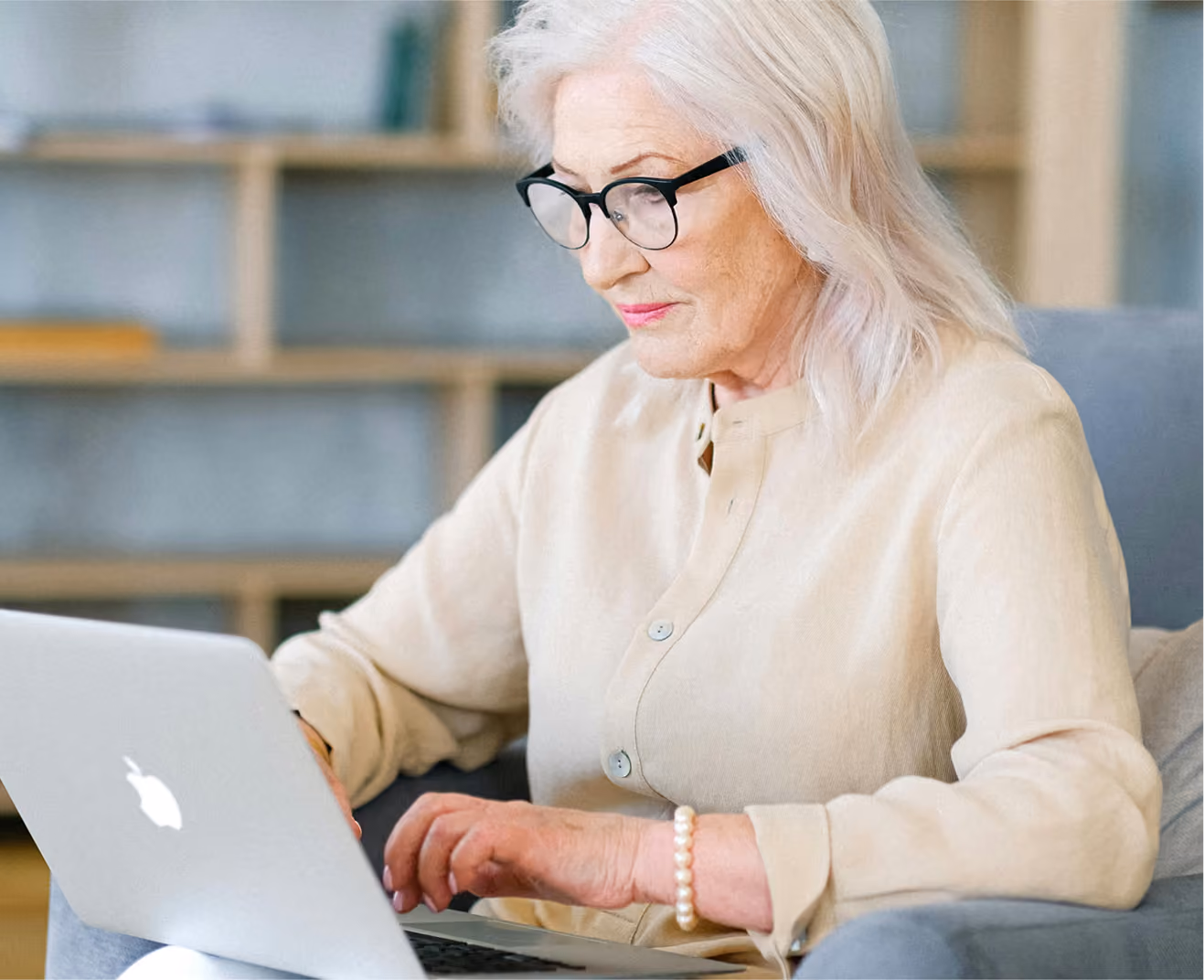 Elderly woman with white hair and glasses working on a laptop while seated in an armchair.