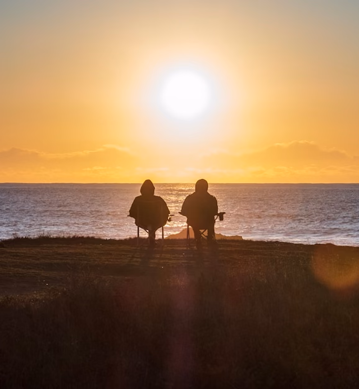 Two people sitting in chairs facing the ocean during a bright sunset.