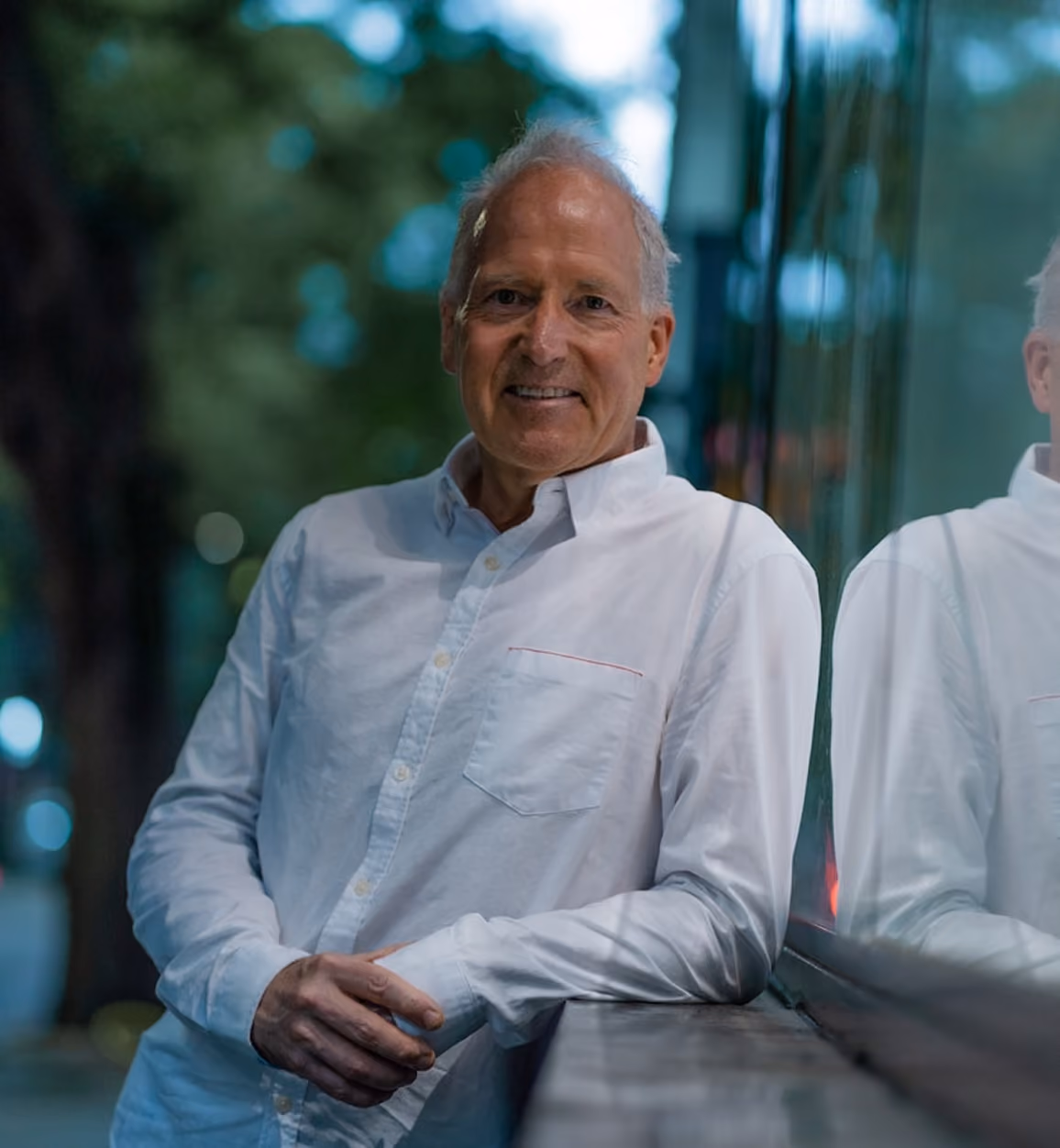 Older man in white shirt leaning on a glass wall, smiling with blurred trees in the background.