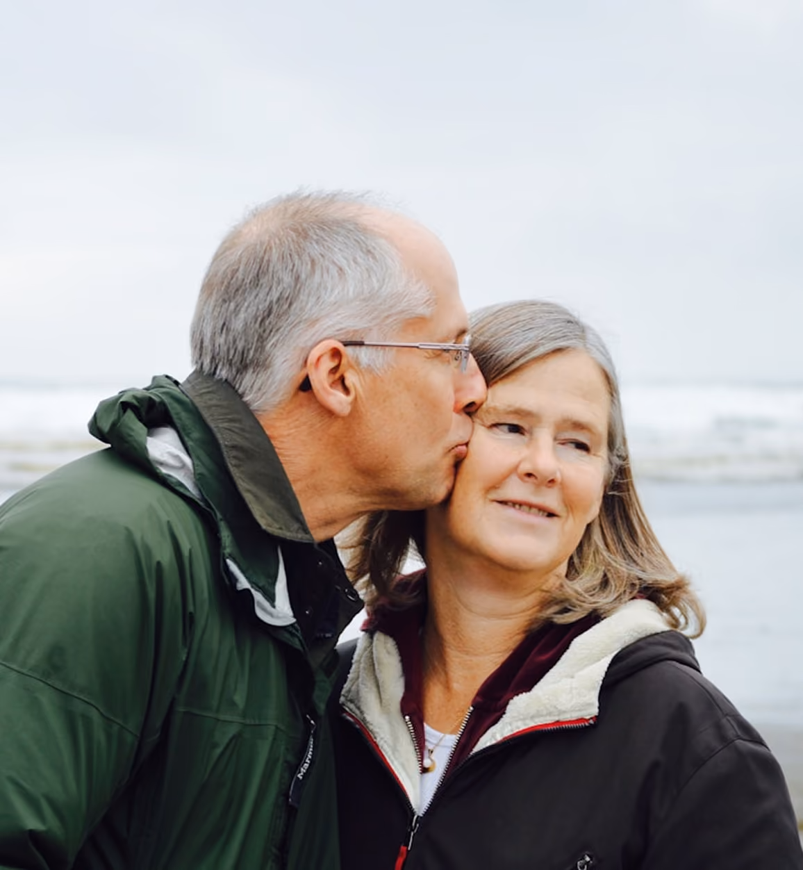 Older man in green jacket kissing older woman in black jacket on the cheek by the sea.