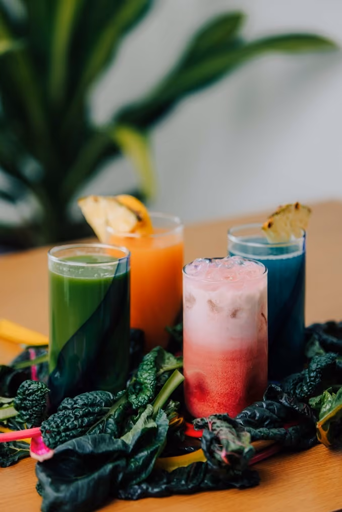 Four colorful juice drinks surrounded by fresh leafy greens and topped with pineapple slices.