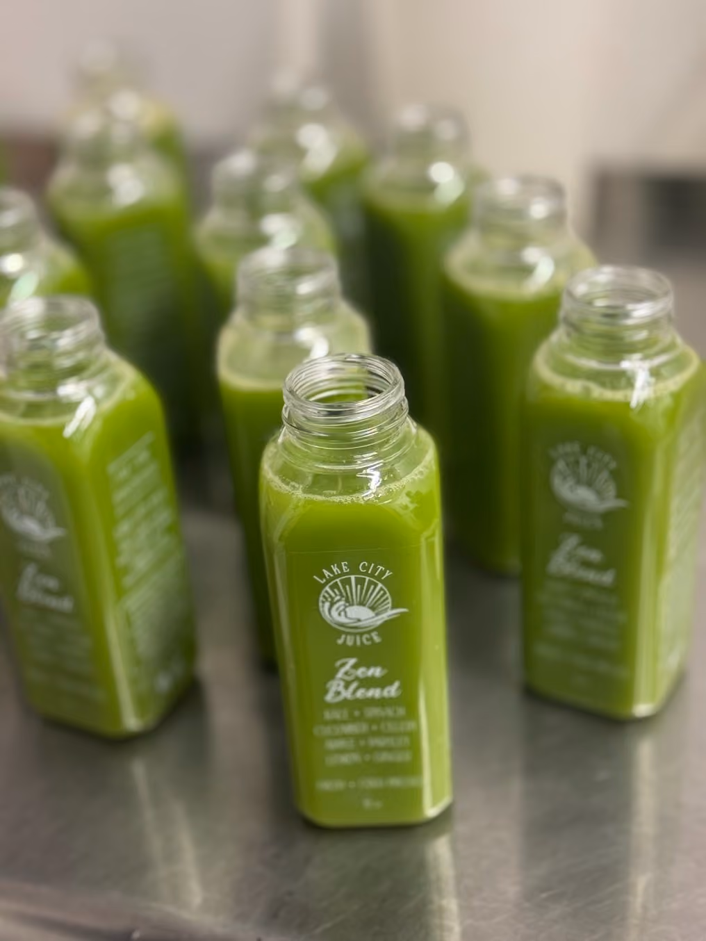 Several clear bottles of green juice labeled Lake City Juice Zen Blend on a metal surface.