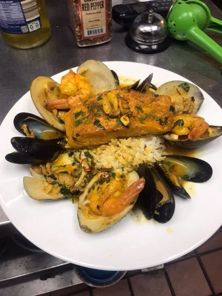Plate with cooked salmon, mussels, shrimp, clams, and rice garnished with herbs and sauce.