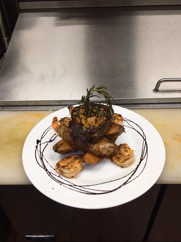 Grilled steak with garlic and rosemary on roasted potatoes, shrimp, and balsamic glaze on white plate.