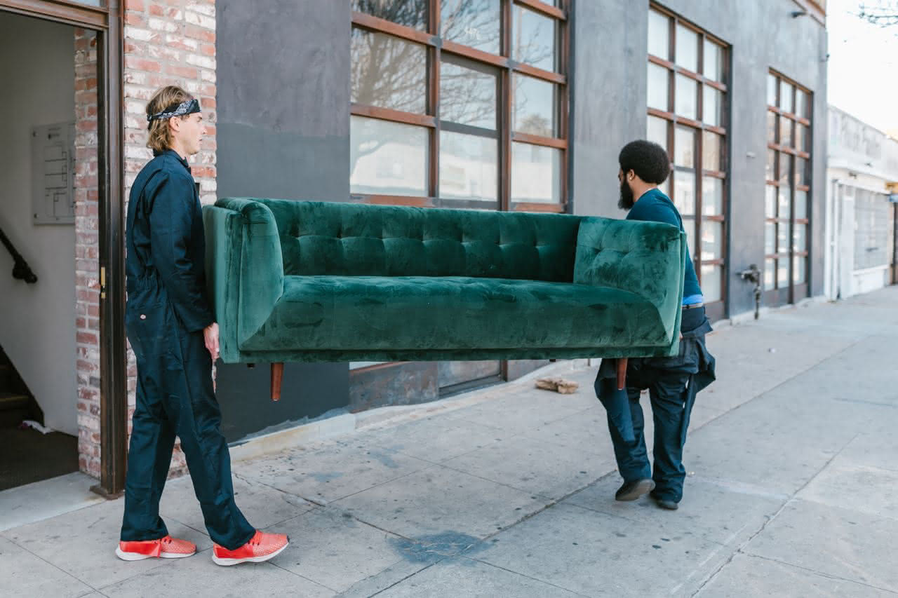 Zwei Männer tragen ein grünes Sofa auf einem Bürgersteig neben einem Gebäude mit großen Fenstern.