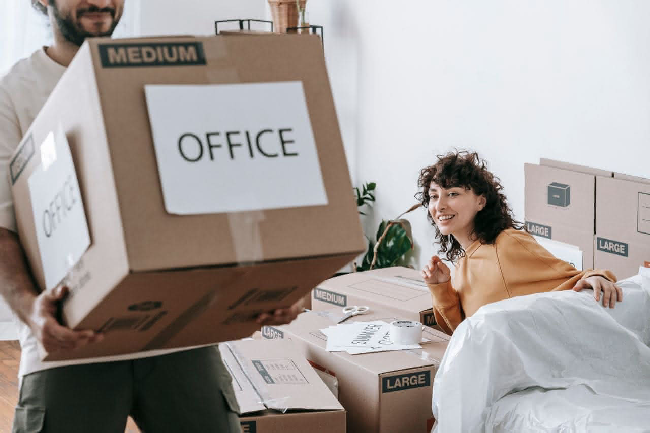 Mann trägt Karton beschriftet mit Büro, während Frau neben weiteren Umzugskartons lächelnd sitzt.