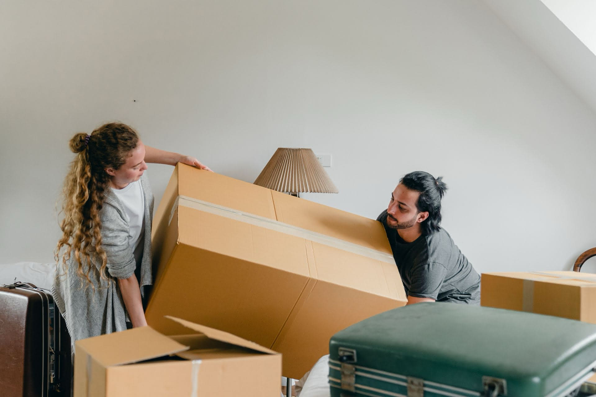 Zwei Personen heben zusammen einen großen Karton in einem Zimmer mit weiteren Kisten und Koffern.