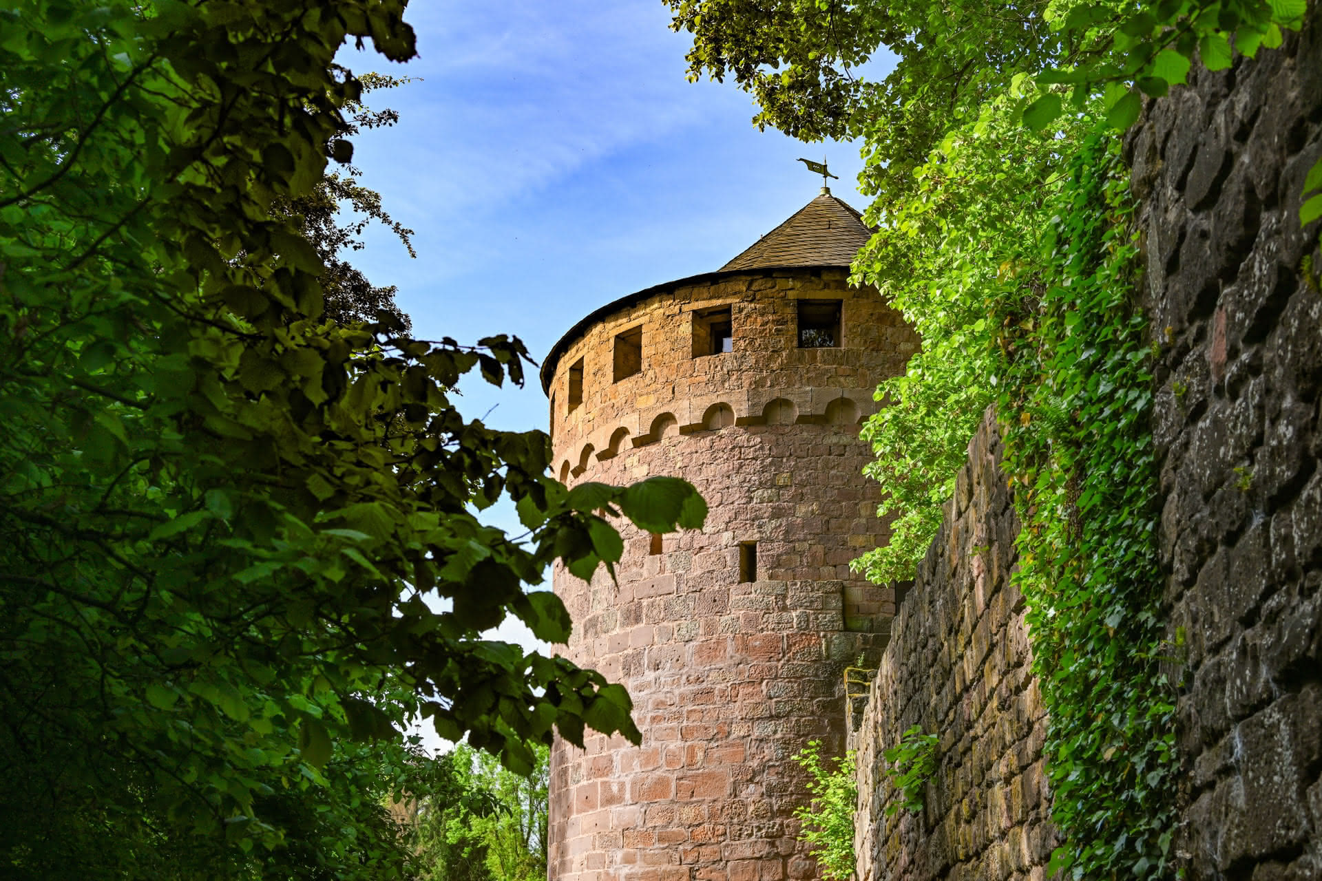 Rundturm aus Stein mit Zinnen, umgeben von grünen Bäumen und einer Steinmauer.
