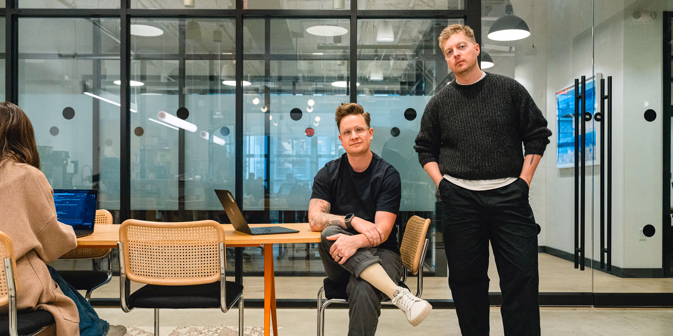 Benten Woodring and Devin Fountain posing in an office. Benten is sitting crossed legged on a modern wicker chair. Devin is standing with his hands in his pockets. Victoria is just off camera to the left working on her laptop.