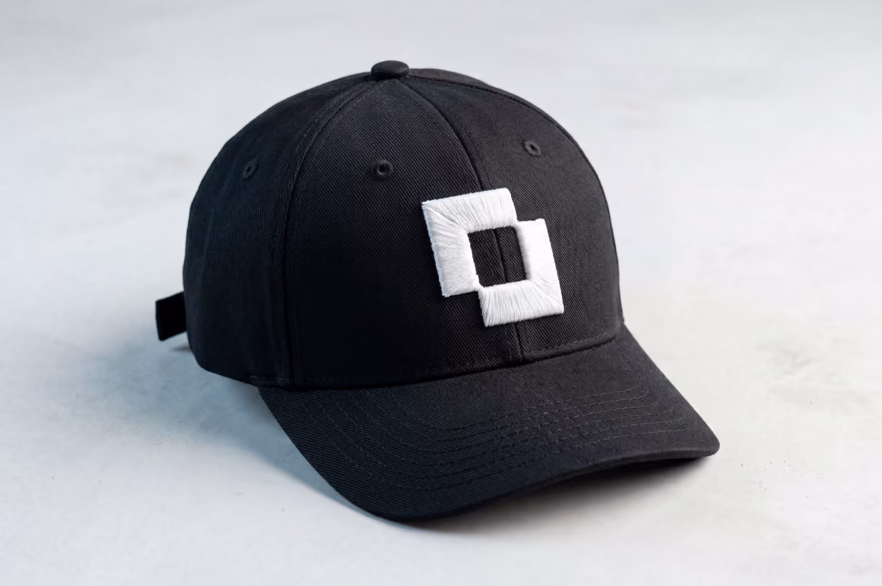 Black baseball cap embroidered with a white square logo, photographed against a soft gray background.