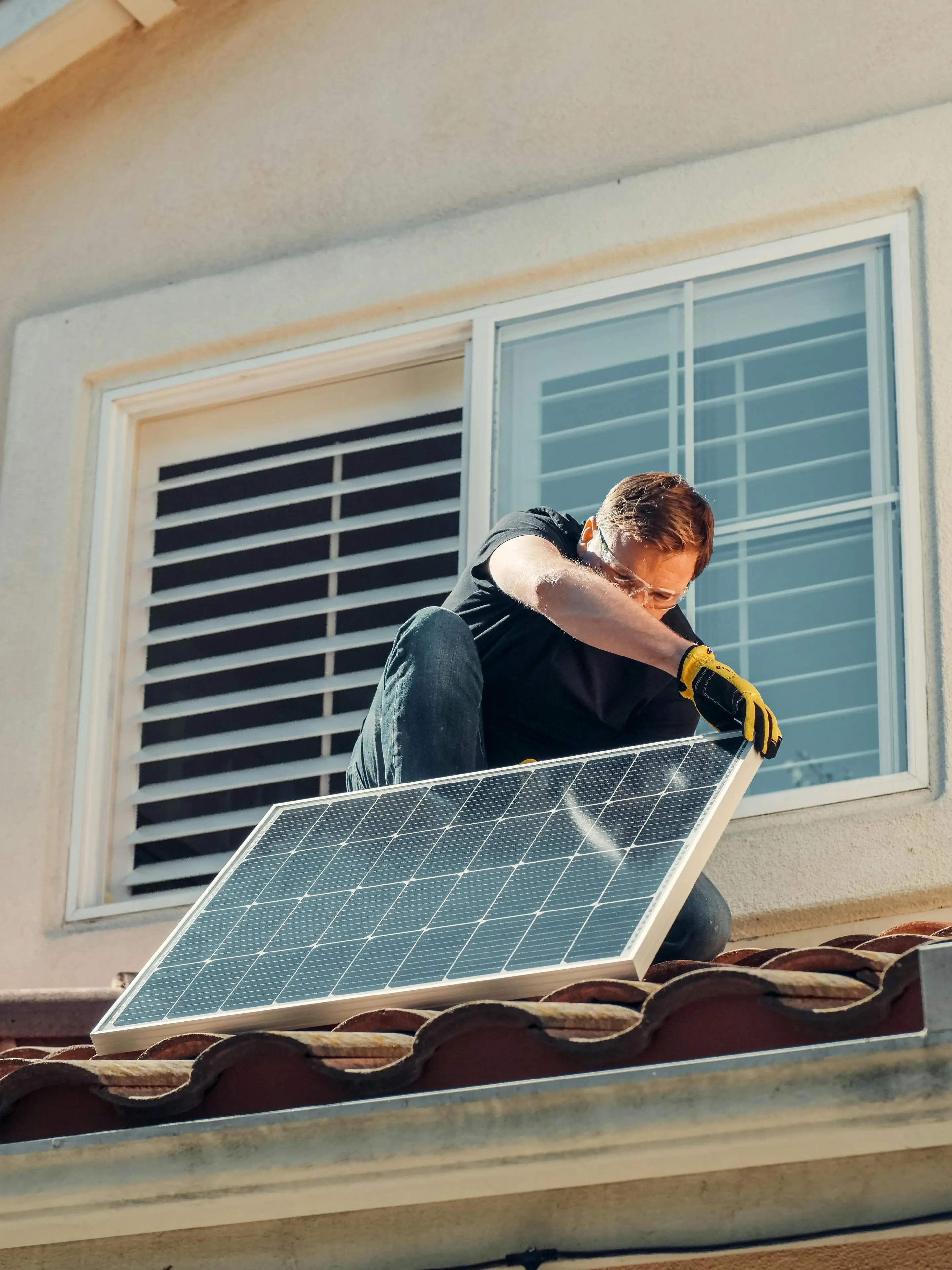 Person wearing gloves installing a solar panel on a rooftop near a window.