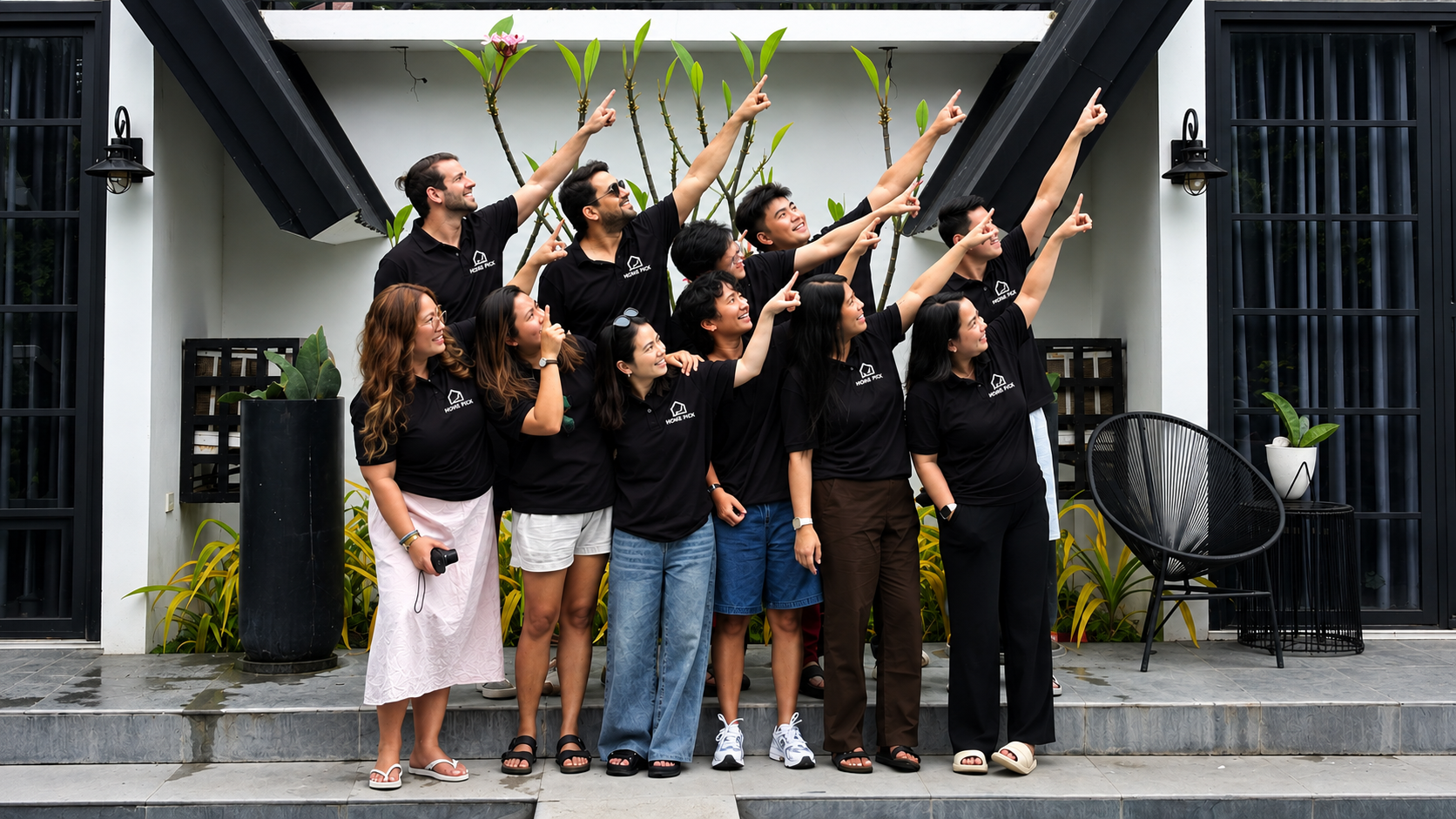Group of eleven people standing on steps in front of a building with plants, all wearing black shirts with a white logo, smiling and pointing upwards and to the right.