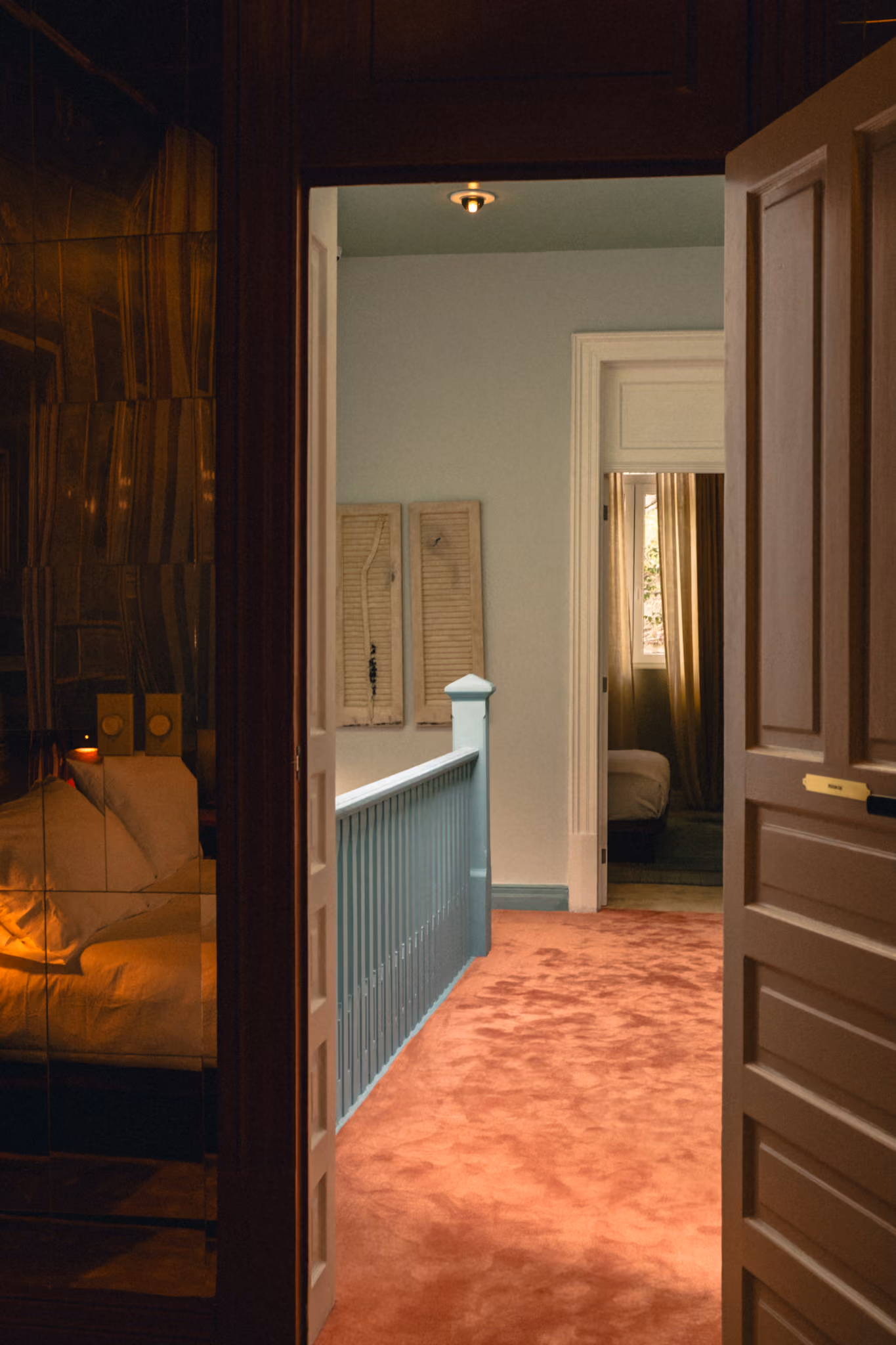 View through open doors of a hallway with dusty rose carpet and a teal railing leading to a dimly lit bedroom.