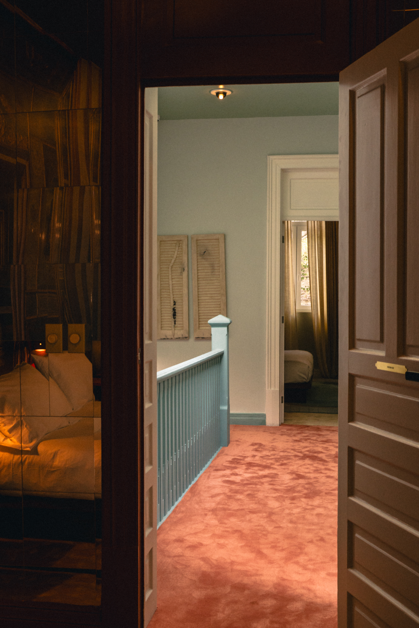 View through open doors of a hallway with dusty rose carpet and a teal railing leading to a dimly lit bedroom.
