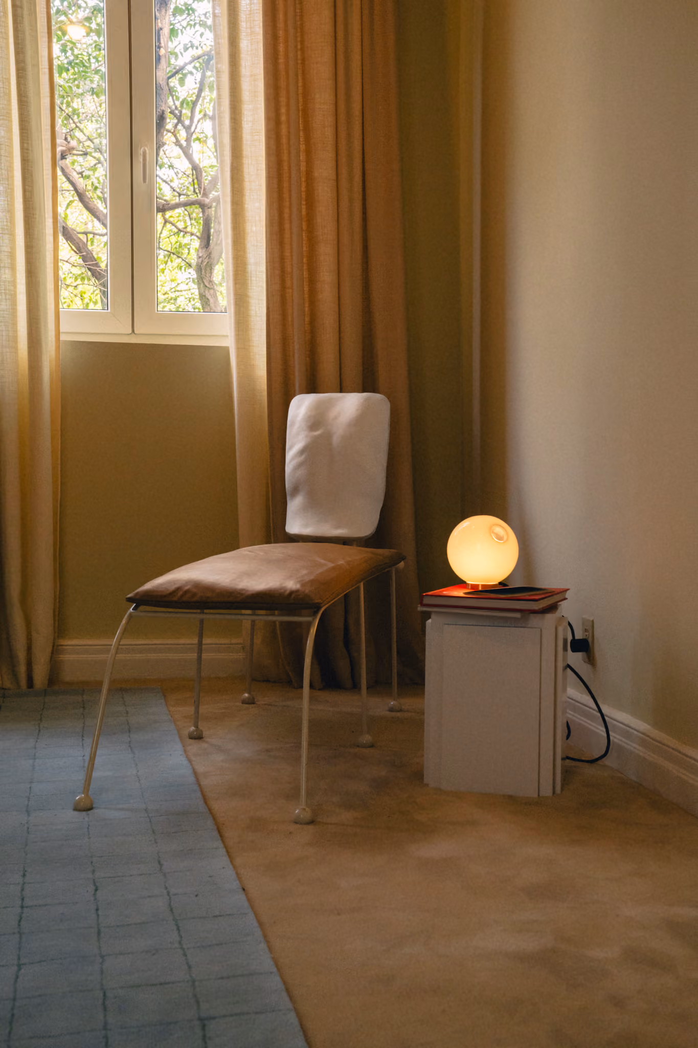 Modern chair with white backrest beside a white nightstand holding a glowing round lamp and books.