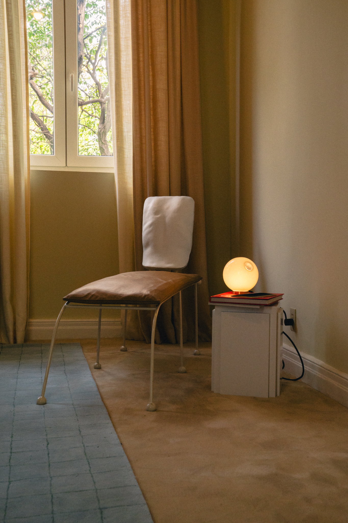 Modern chair with white backrest beside a white nightstand holding a glowing round lamp and books.