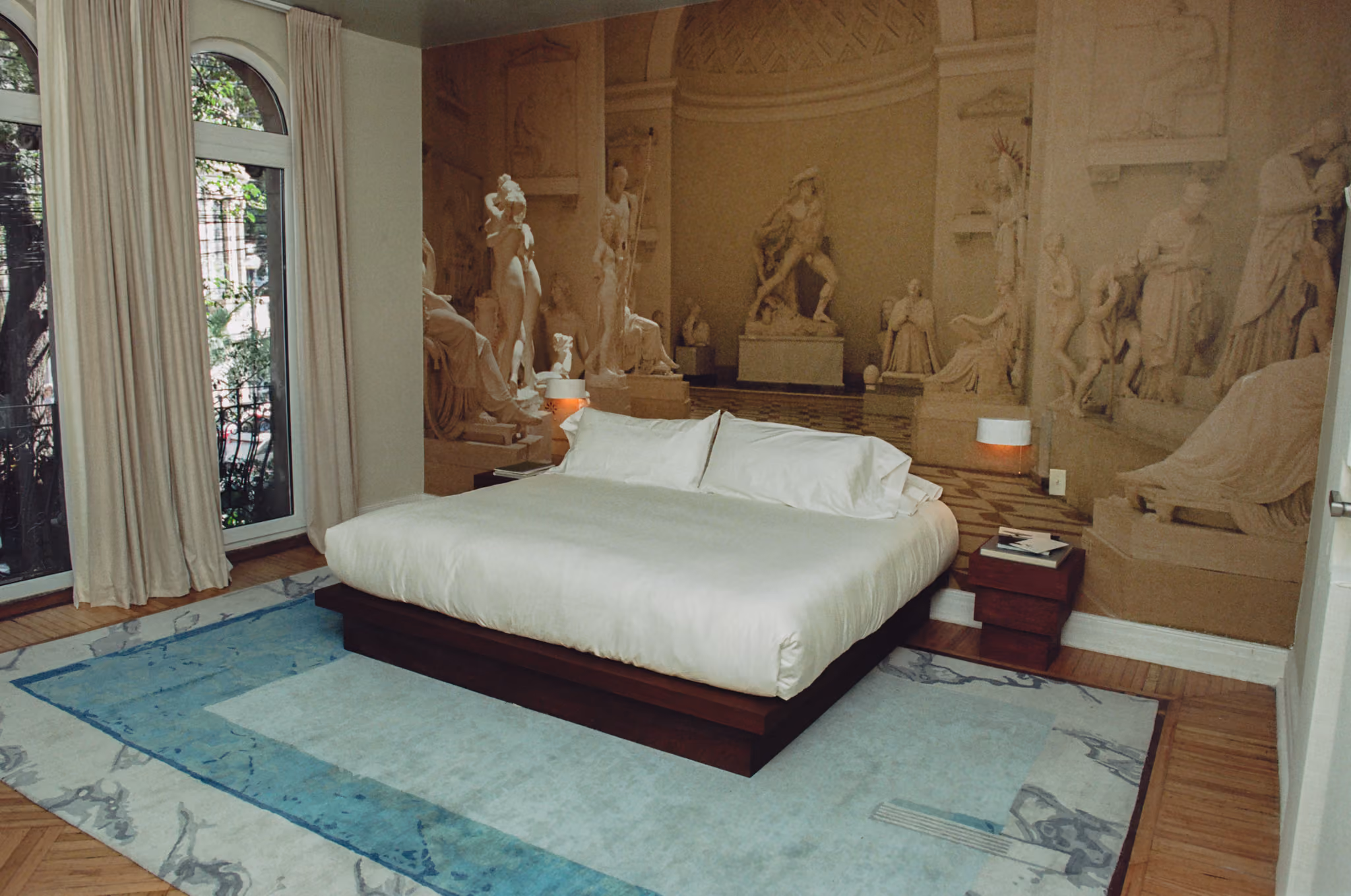 Modern bed with white bedding on blue rug in room with classical statue mural wall and arched window.