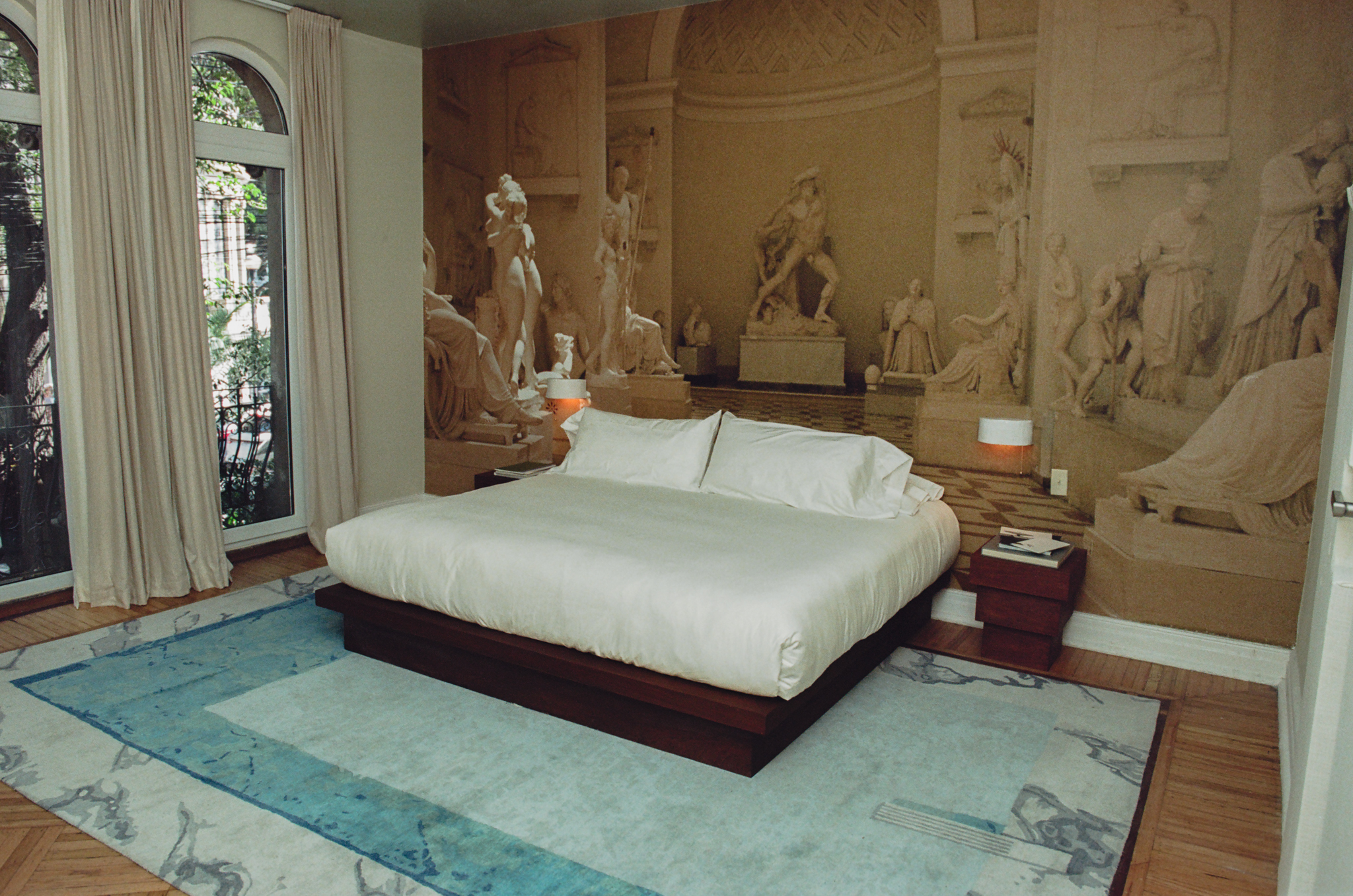 Modern bed with white bedding on blue rug in room with classical statue mural wall and arched window.