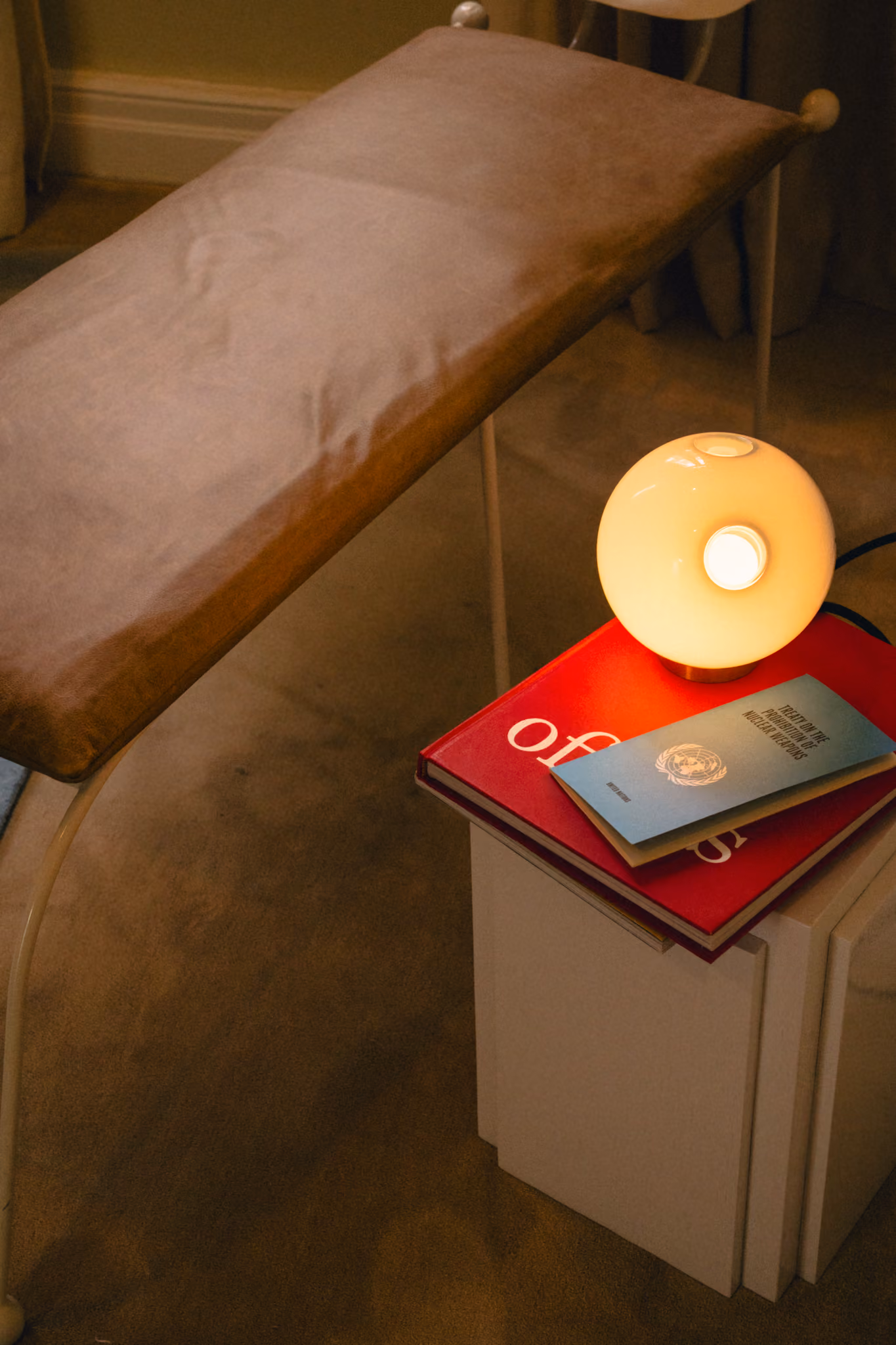 Brown padded bench near a white nightstand with a round lamp and two books on top.