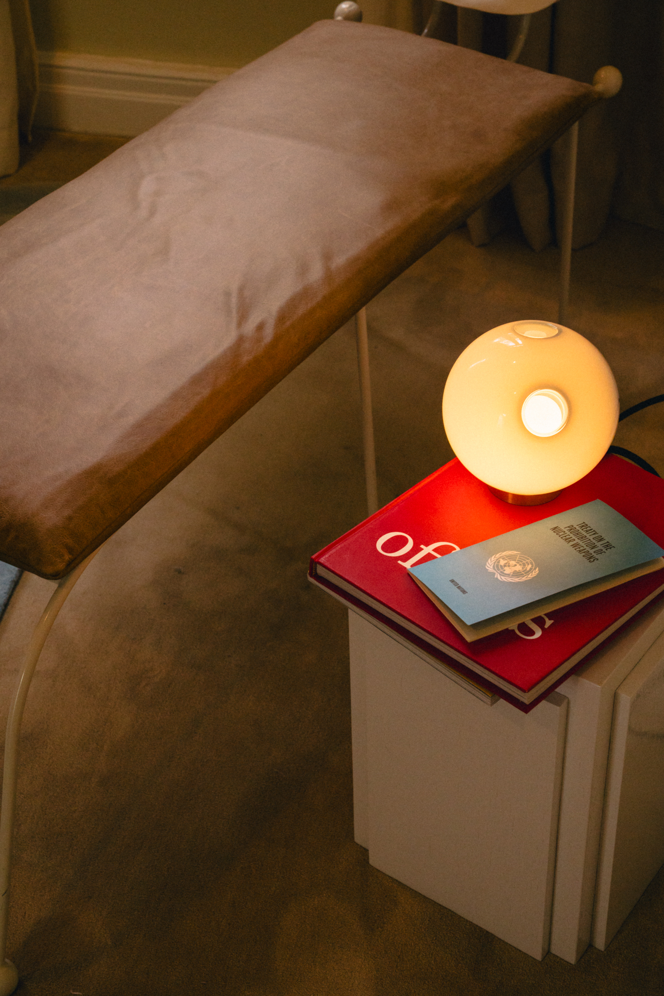 Brown padded bench near a white nightstand with a round lamp and two books on top.