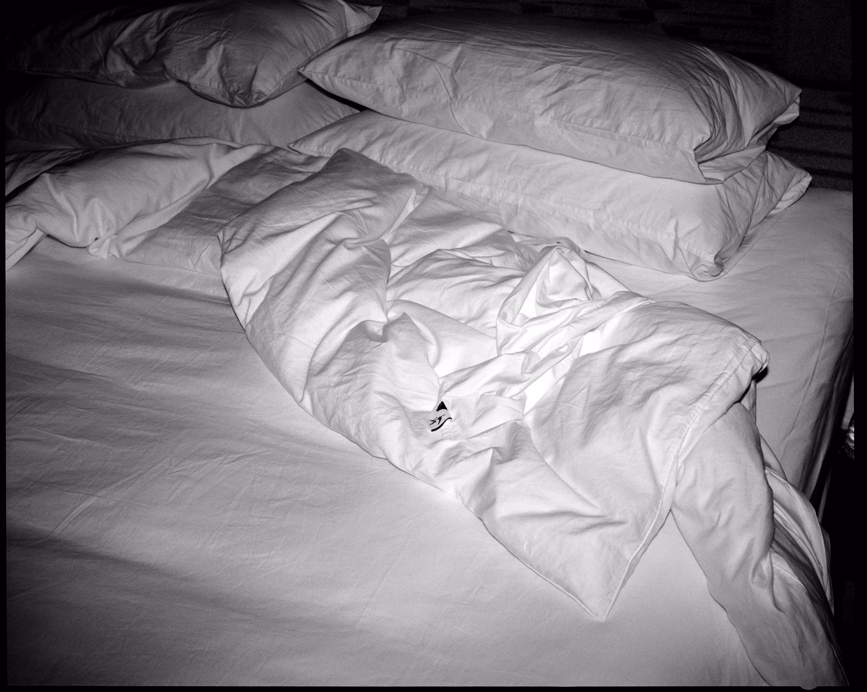 Unmade bed with white crumpled sheets and four stacked pillows.