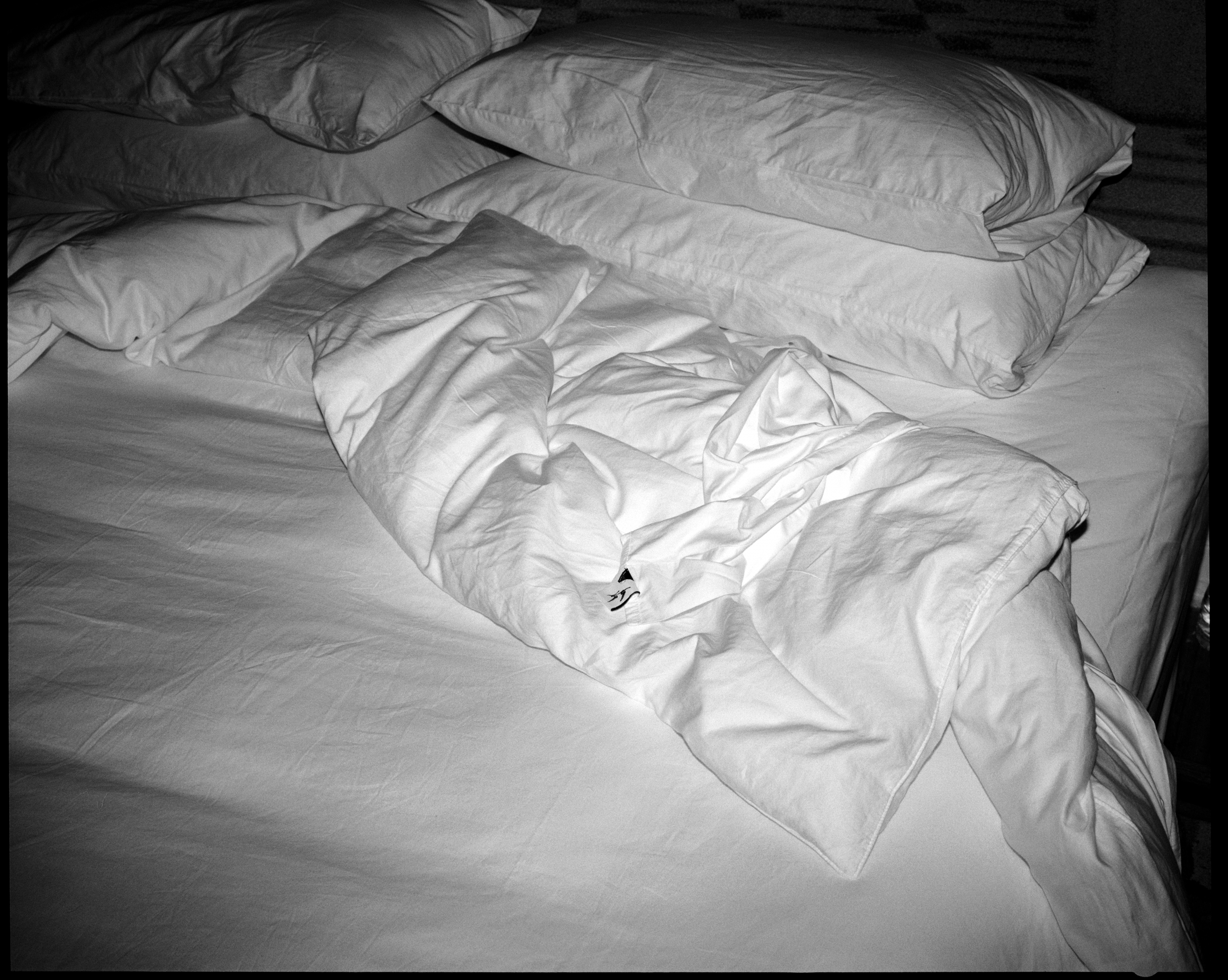 Unmade bed with white crumpled sheets and four stacked pillows.
