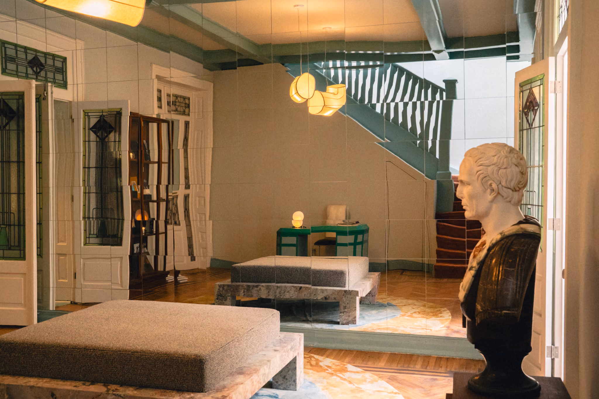 Room with a marble bust, mirrored wall, staircase, modern lighting, and cushioned benches.