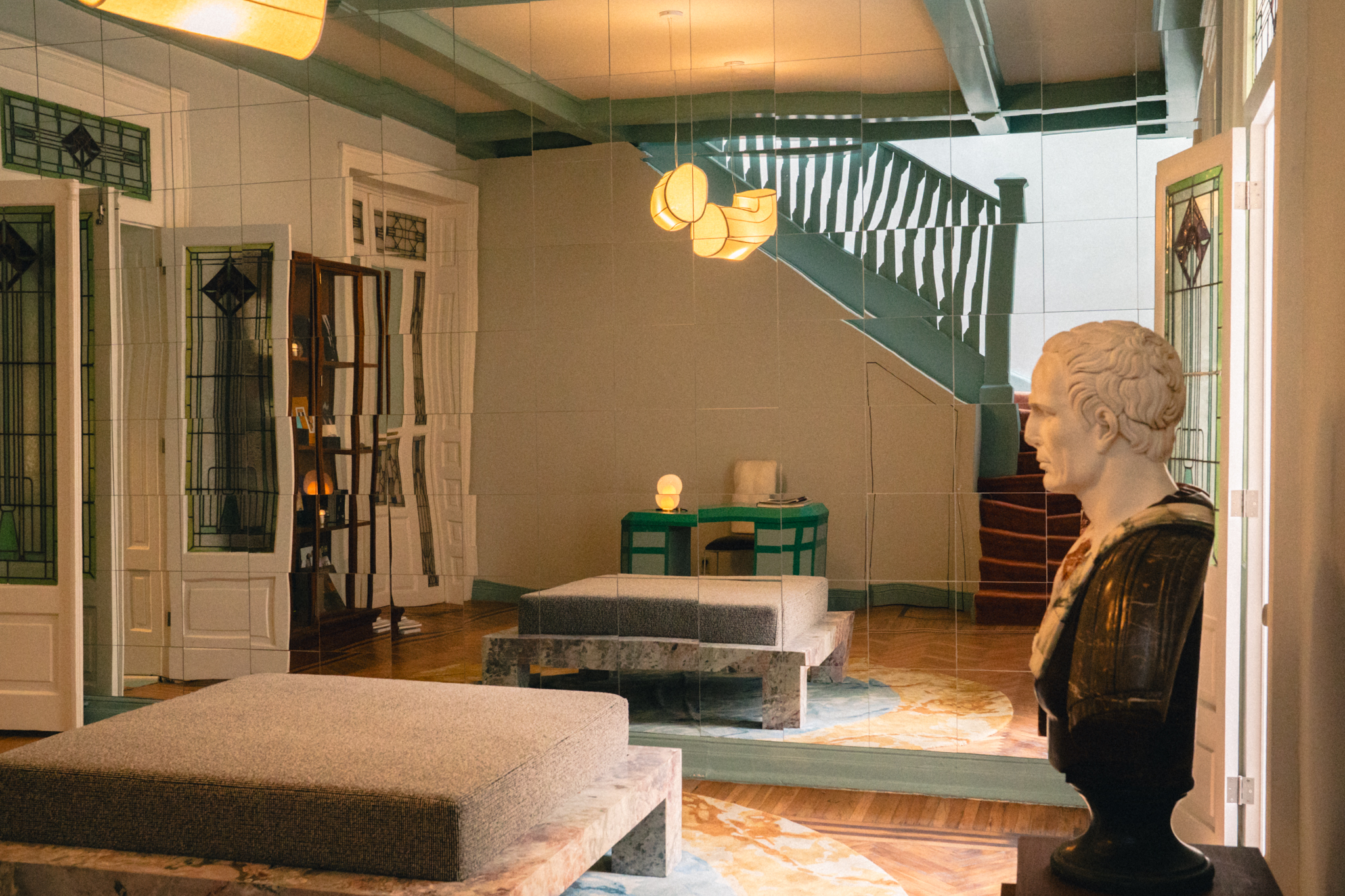 Room with a marble bust, mirrored wall, staircase, modern lighting, and cushioned benches.