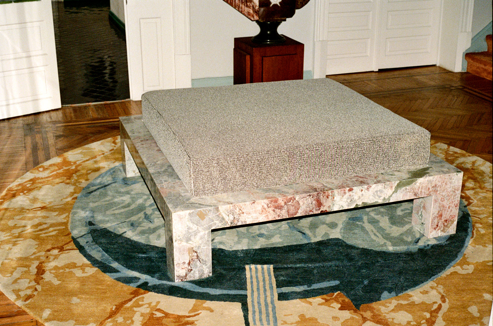 Square marble table with a large gray cushion on top, placed on a round multicolored rug.