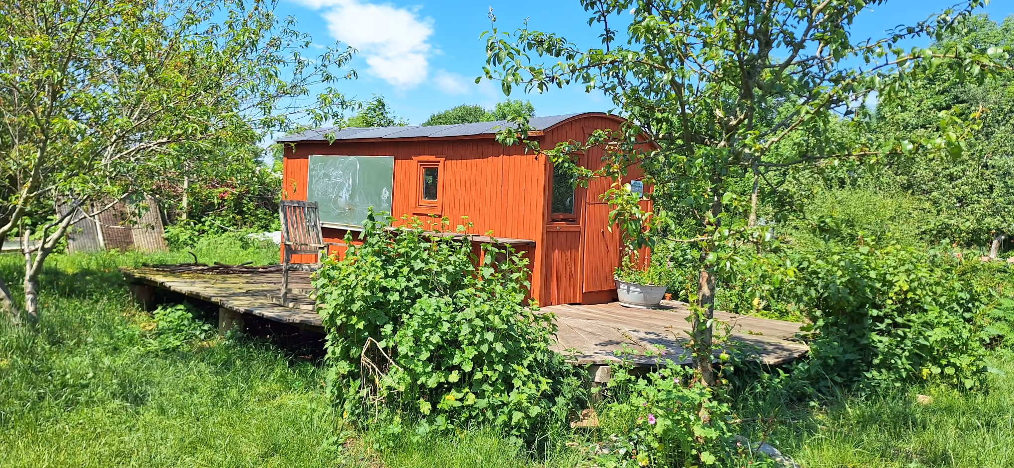 Rotes Gartenhaus mit Holzveranda, Stuhl und grünen Büschen unter blauem Himmel.