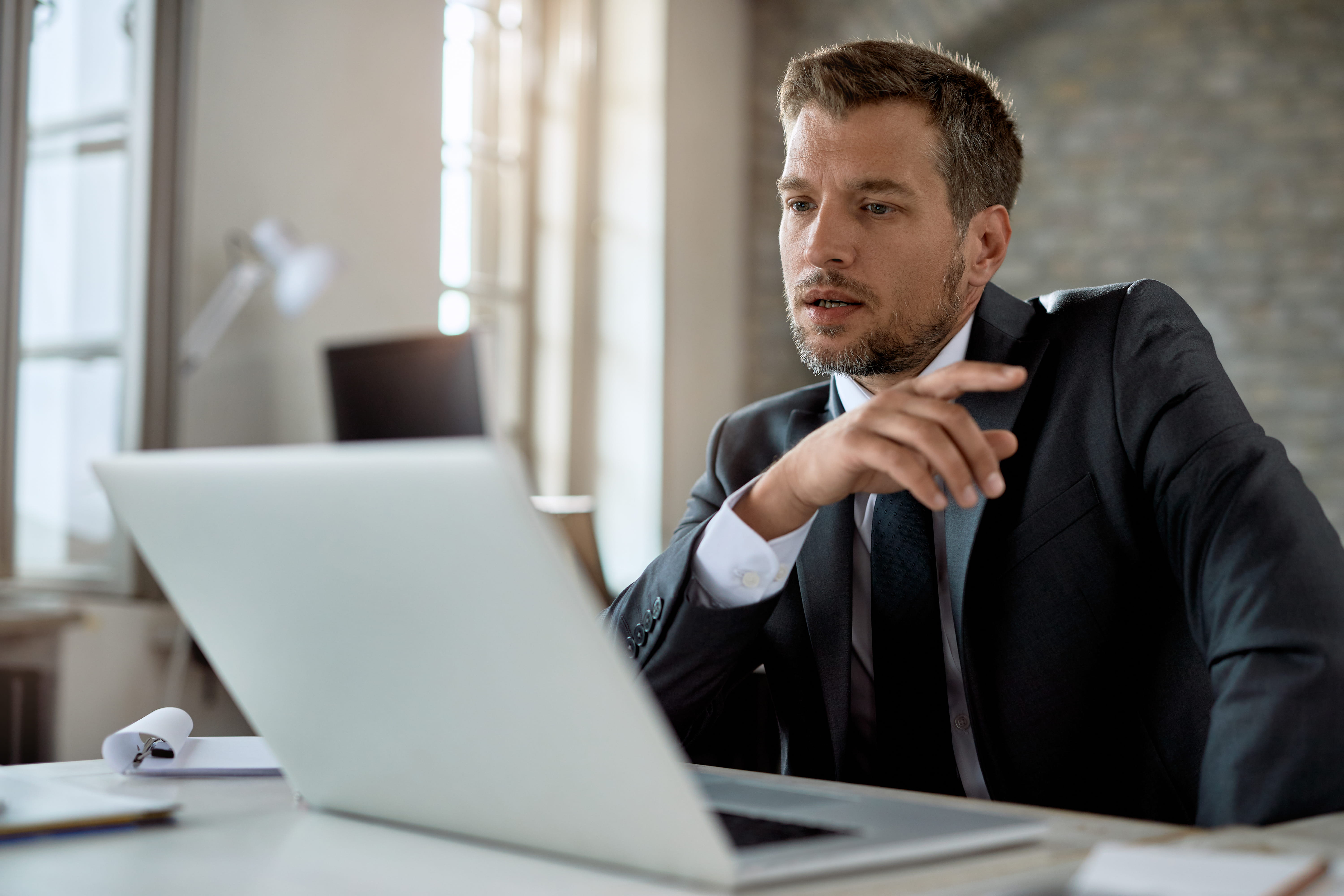 Pensive businessman in a dark suit sitting at a desk, looking at a laptop with a thoughtful expression in a softly lit office.