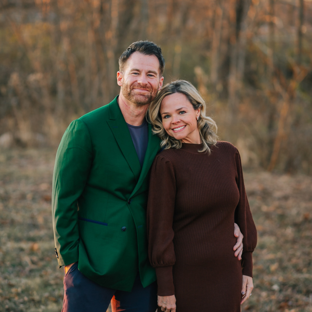 Man in green blazer and woman in brown dress standing closely outdoors in autumn setting, smiling.