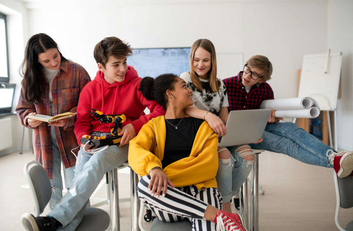 Group of five diverse teenagers studying together with laptop, robot arm, book, and blueprints in classroom.