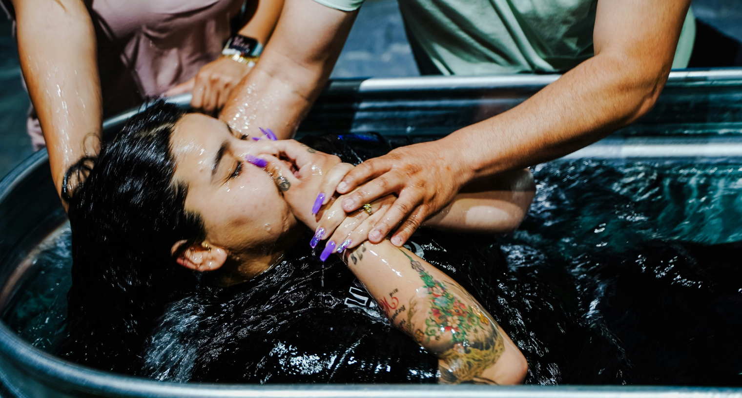 Person being baptized in a water tub with hands covering their nose, assisted by others.
