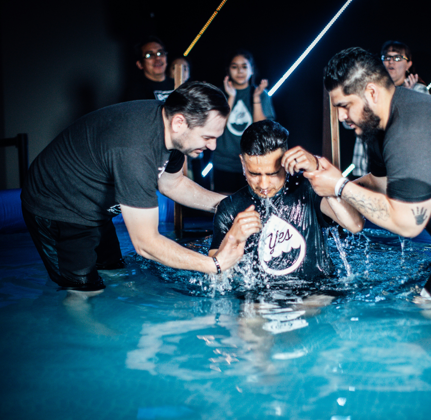 Man being baptized in a pool with others supporting him, water splashing around.