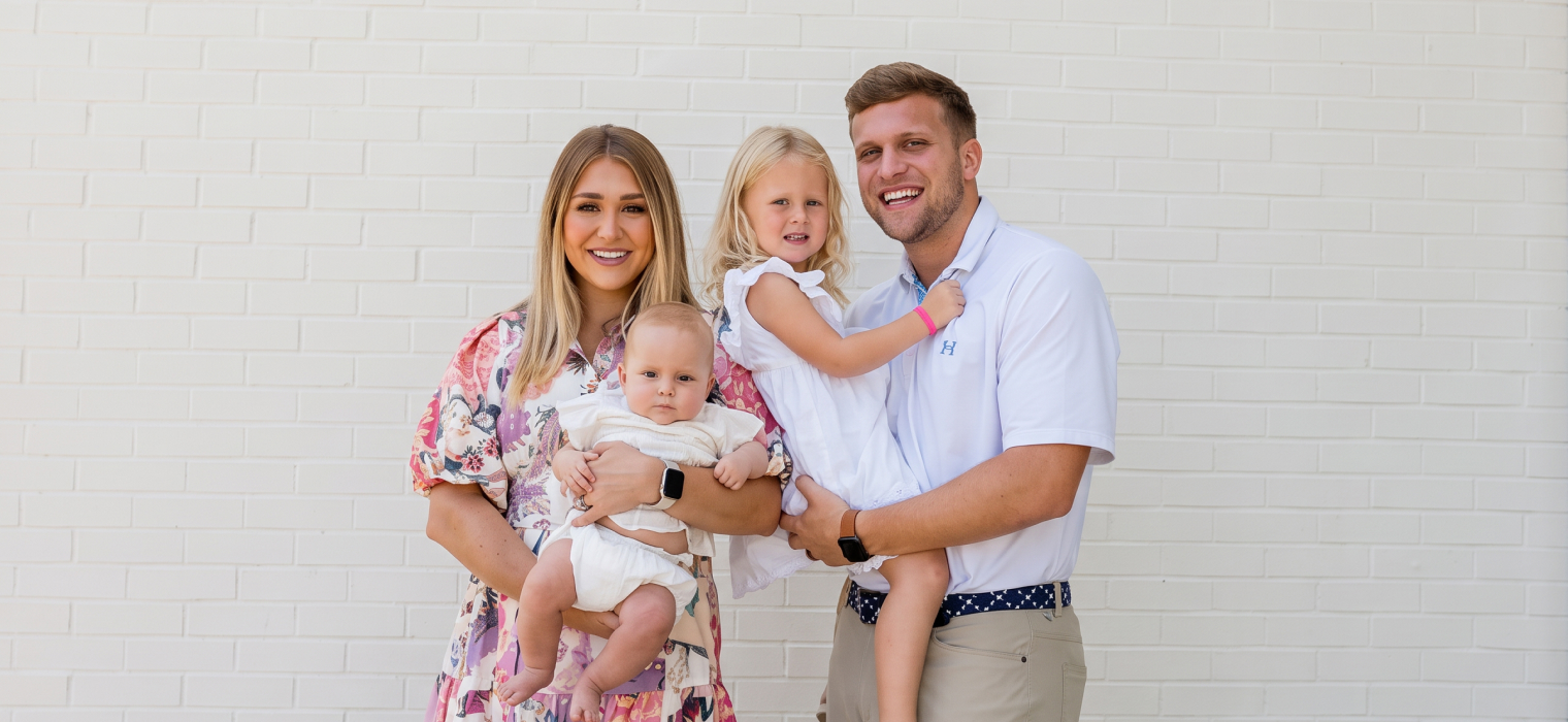 Smiling couple holding two young children, standing in front of a white brick wall.