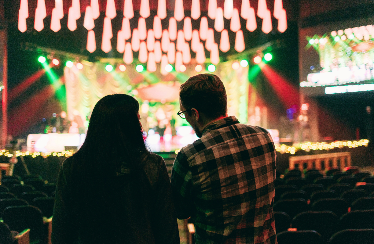 Two people viewed from behind sitting in a dim auditorium with a bright stage and hanging lights.