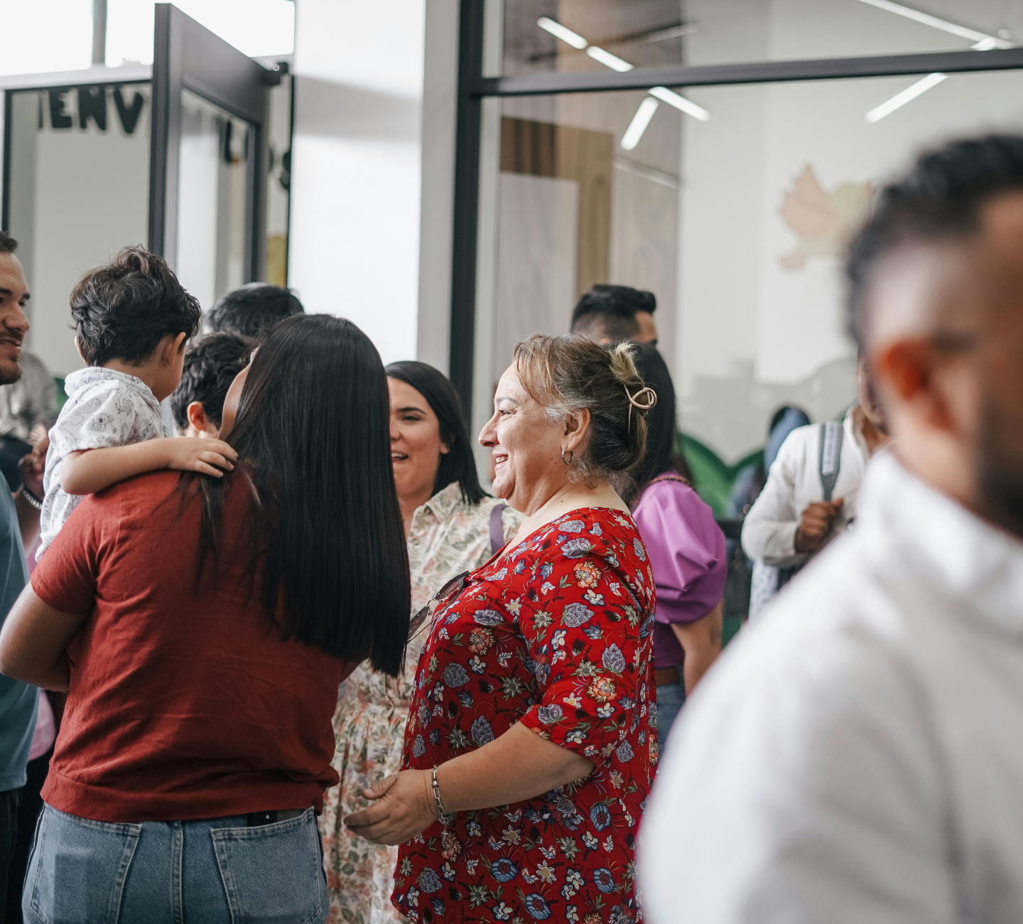People socially interacting indoors, including a woman in a red floral blouse smiling and a child being held.