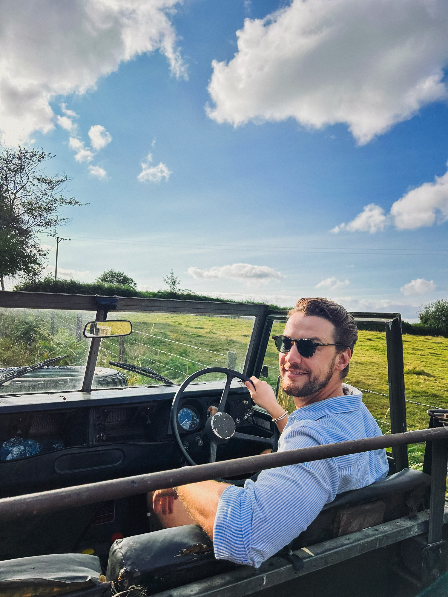 Man wearing sunglasses and a light blue striped shirt driving an open-top vintage off-road vehicle in a sunny countryside with green grass and blue sky with clouds.